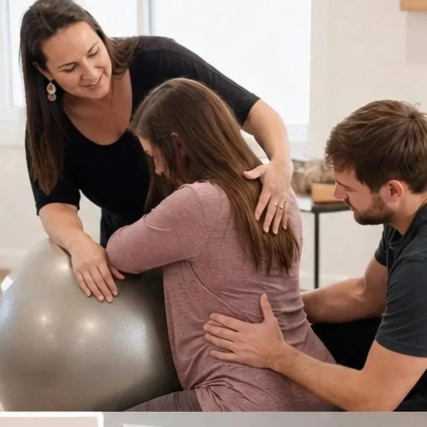A woman appears to be giving birth with support from a man and a woman at a home birth setting, with a large exercise ball involved.