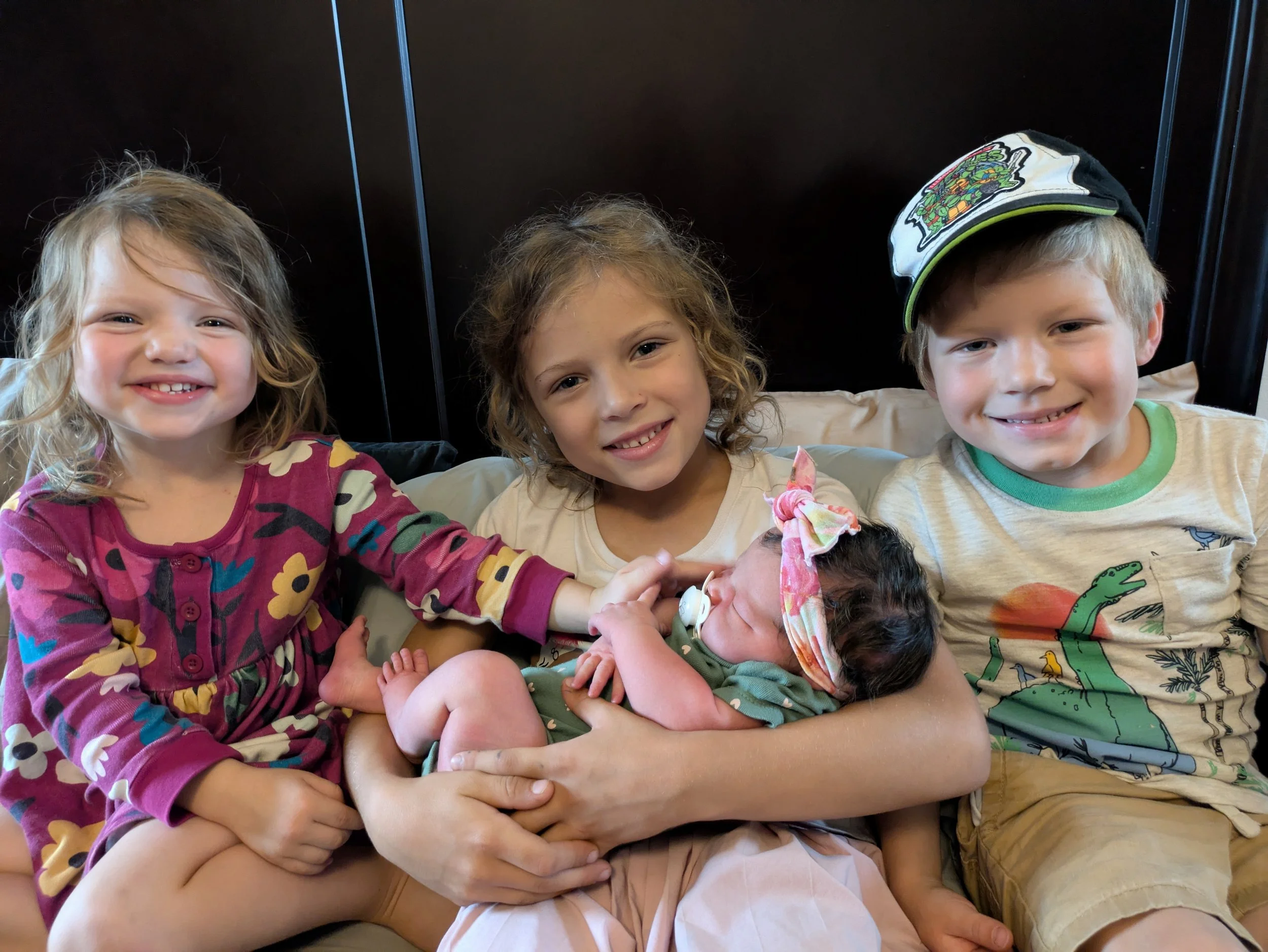 Three children smiling and sitting together, with two of them holding a newborn baby, in an indoor setting.