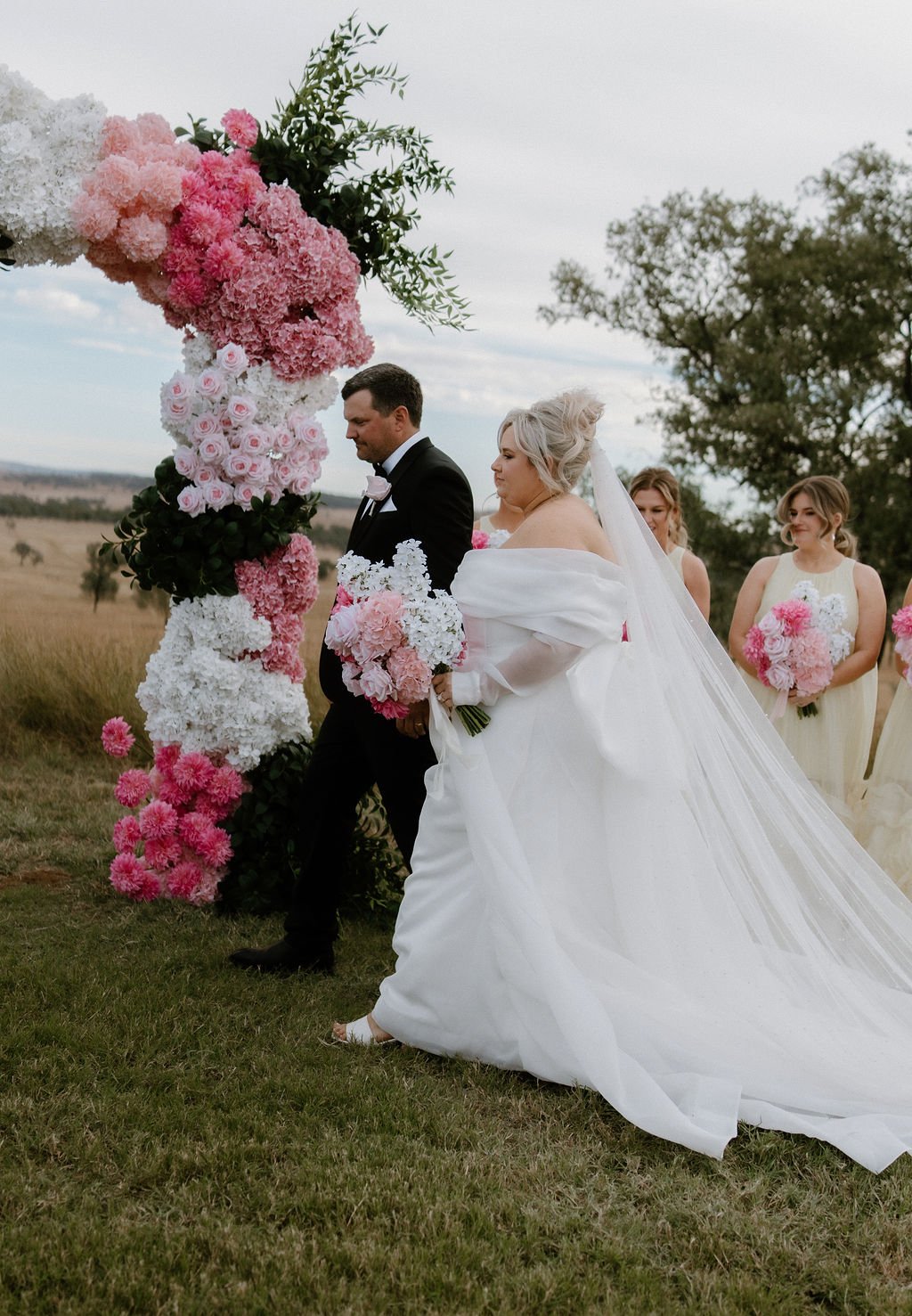 Wedding ceremony outdoors with a bride and groom at an arch decorated with pink, white, and green flowers. The bride is in a white gown with a long train, and the groom is in a black tuxedo. Bridesmaids in light-colored dresses hold bouquets of pink 