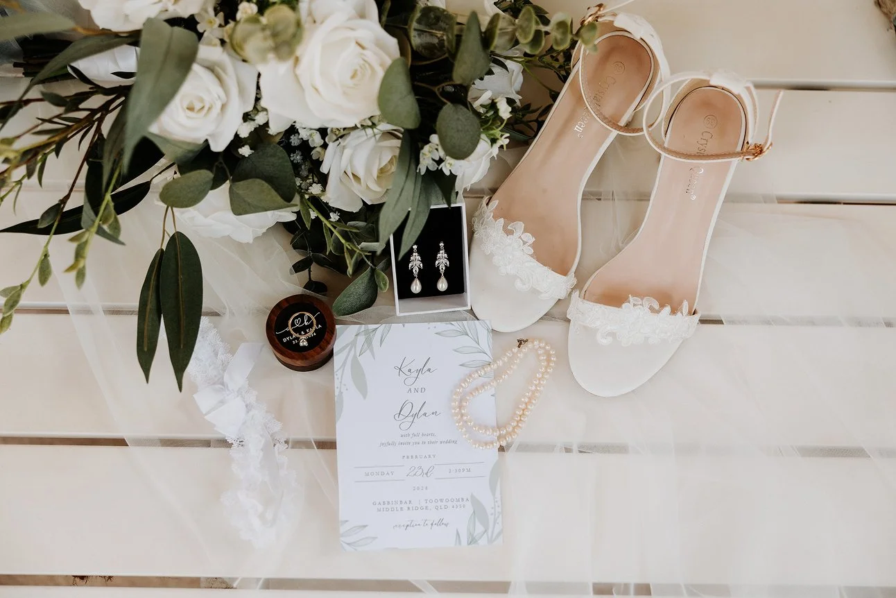 Wedding accessories including white lace heels with ankle straps, pearl jewelry, wedding invitation with green leaf design, pearl necklace, pearl earrings, white lace garter, and a bouquet of white roses and greenery on a white wooden surface.