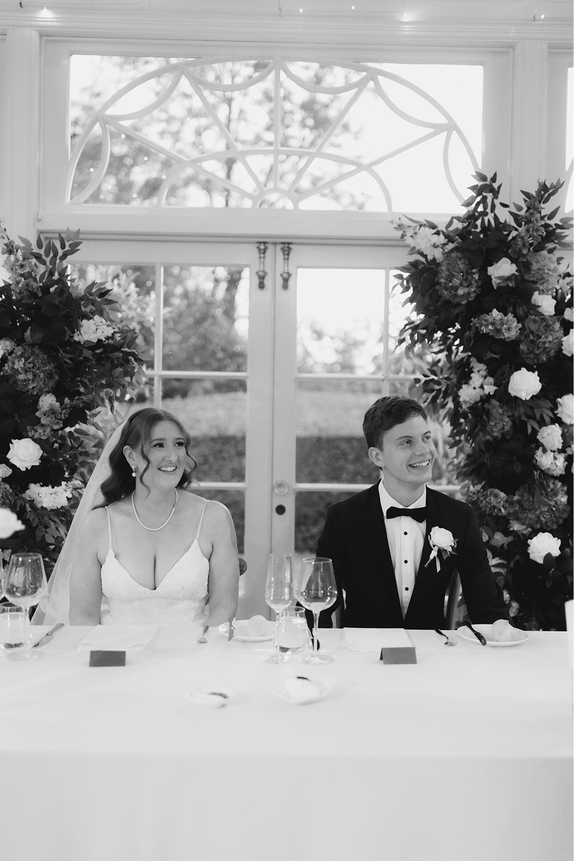 Black and white photo of a bride and groom sitting at a wedding reception table, smiling, with flowers and glassware.