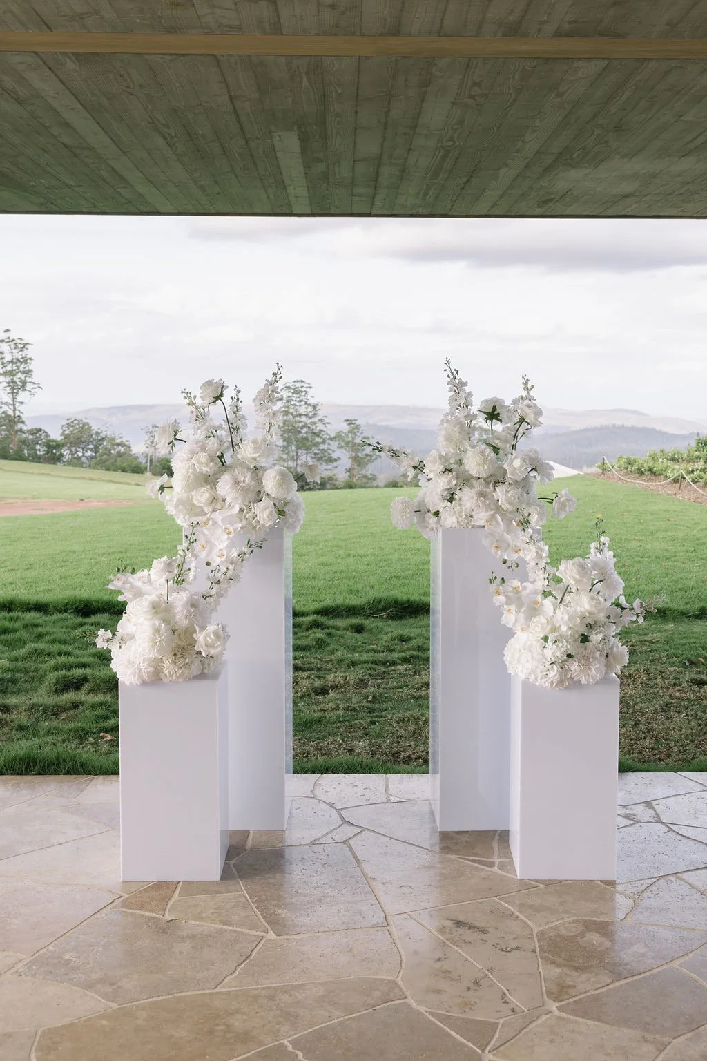 Two white wedding altar stands decorated with white flowers, outdoors with a green lawn and distant hills in the background.