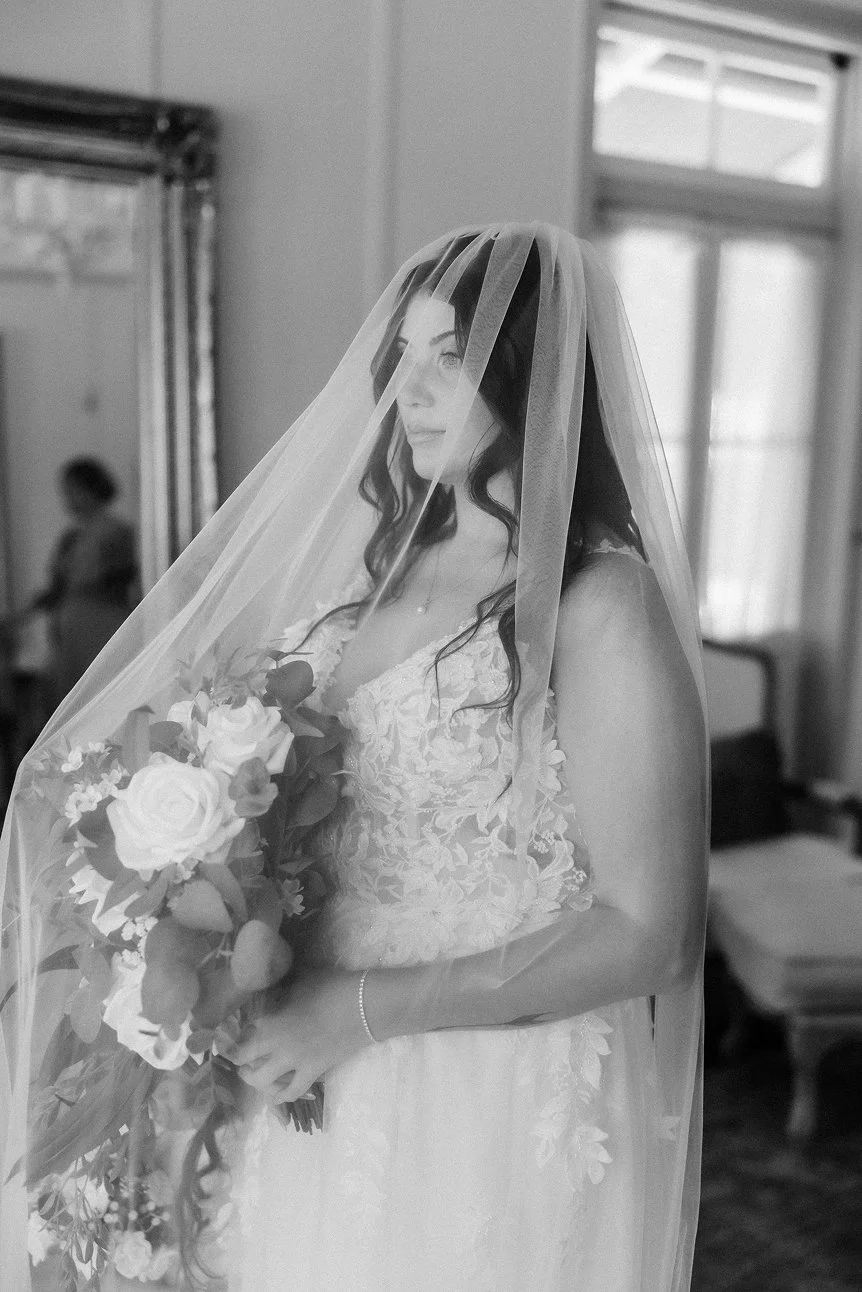 A bride with dark hair in a white lace wedding dress, wearing a veil, holding a bouquet of roses and greenery, indoors near a window.
