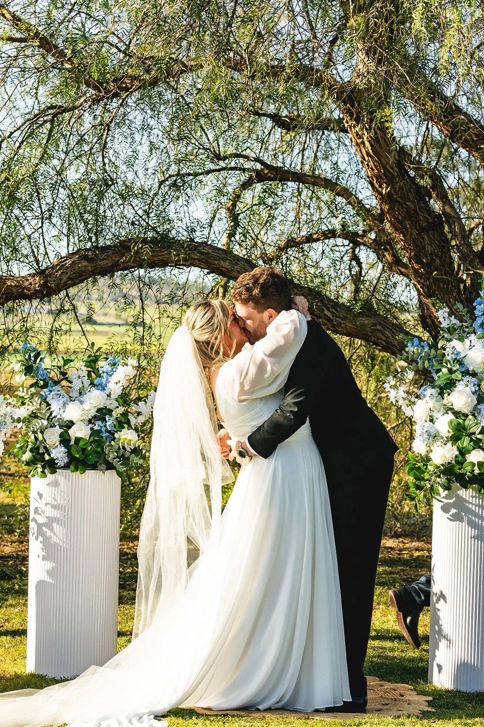 Bride and groom kissing during outdoor wedding ceremony under a tree, with floral arrangements on white pedestals.