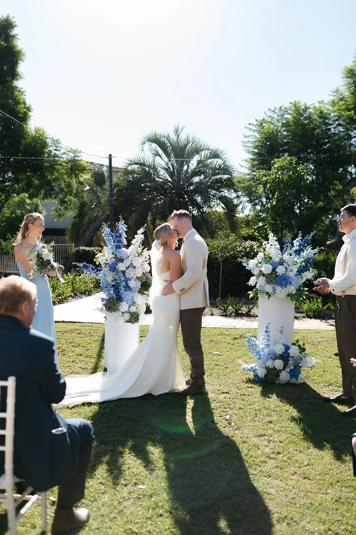 A wedding ceremony taking place outdoors with a bride and groom kissing, surrounded by floral arrangements and guests watching.