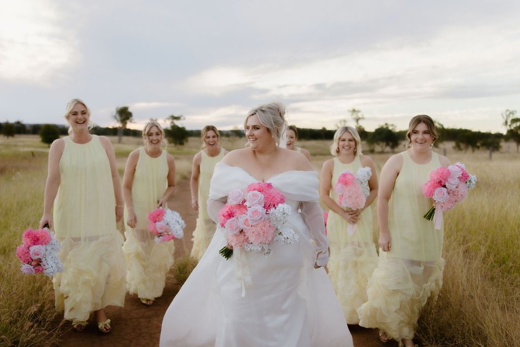 A bride walking with her bridesmaids in a rural field during a wedding photoshoot, holding bouquets of pink and white flowers.
