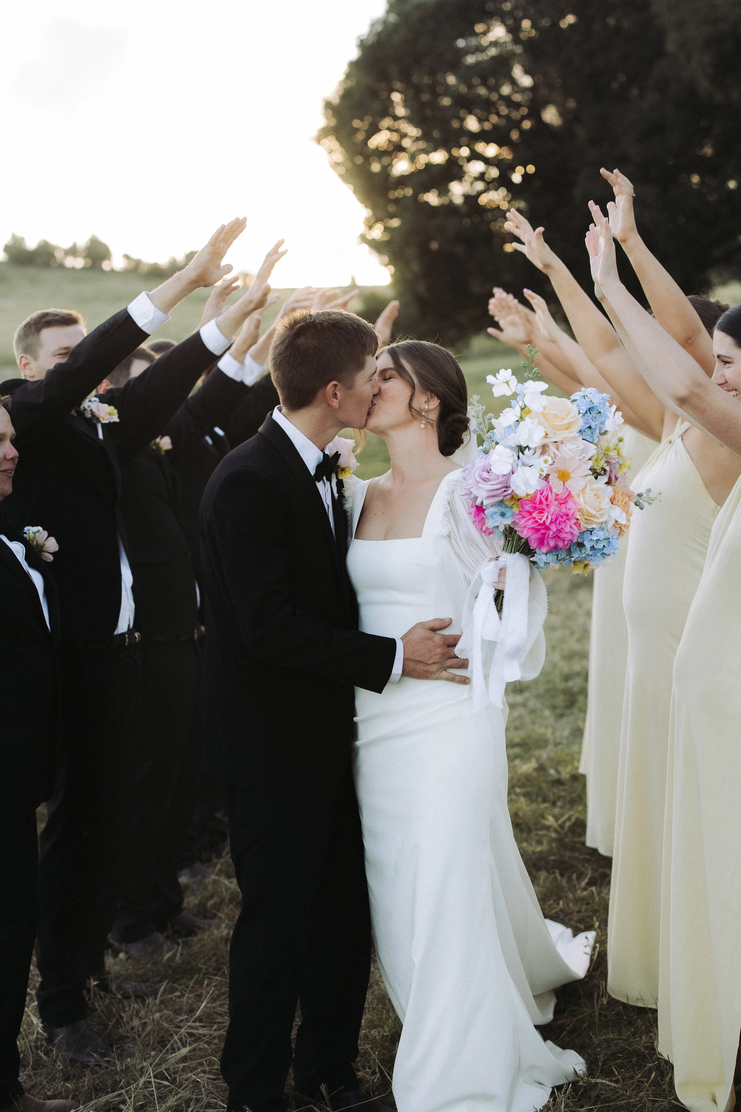 A bride and groom are kissing at their outdoor wedding ceremony, surrounded by bridesmaids and groomsmen raising their hands in celebration.