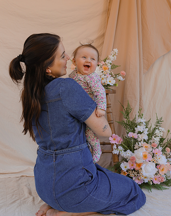 A woman kneeling and holding a baby girl who is smiling and laughing, with a background of floral arrangements and beige curtains.