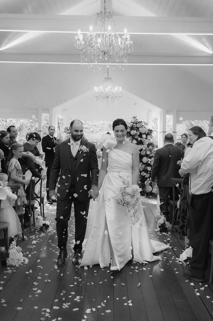 A bride and groom walking down the aisle at their wedding ceremony, surrounded by family and friends, with flower petals falling around them, in a decorated indoor venue with chandeliers.