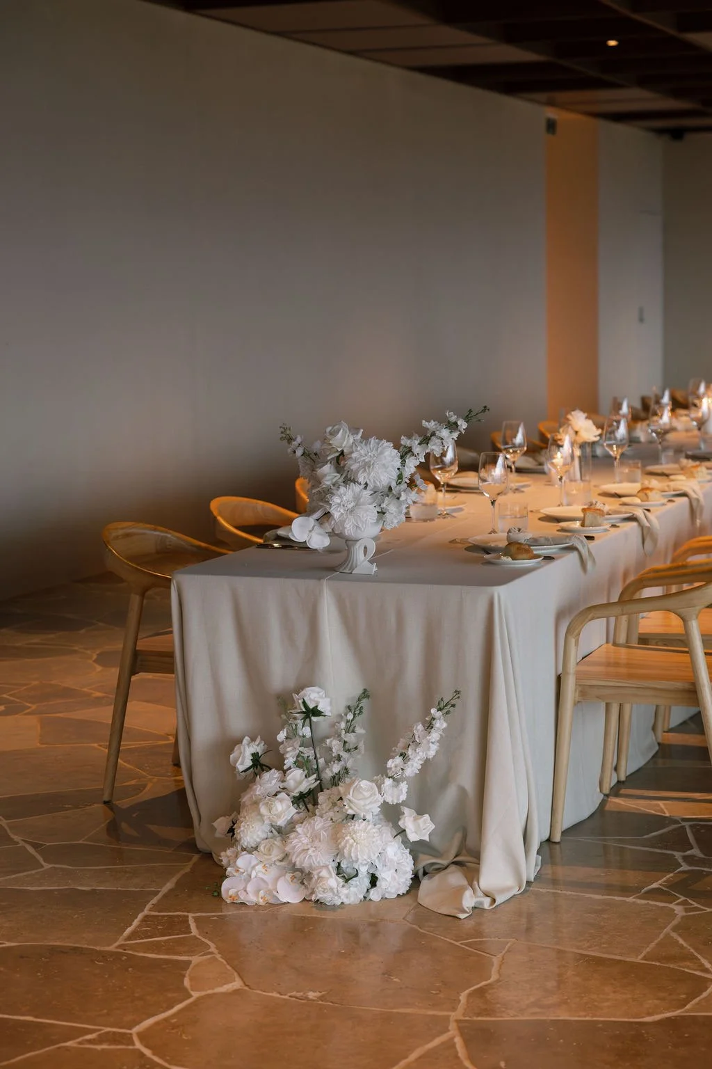 Elegant wedding reception table with white floral centerpiece, wine glasses, plates, and bread rolls, decorated with white flowers and a beige tablecloth, set in a warmly lit room.