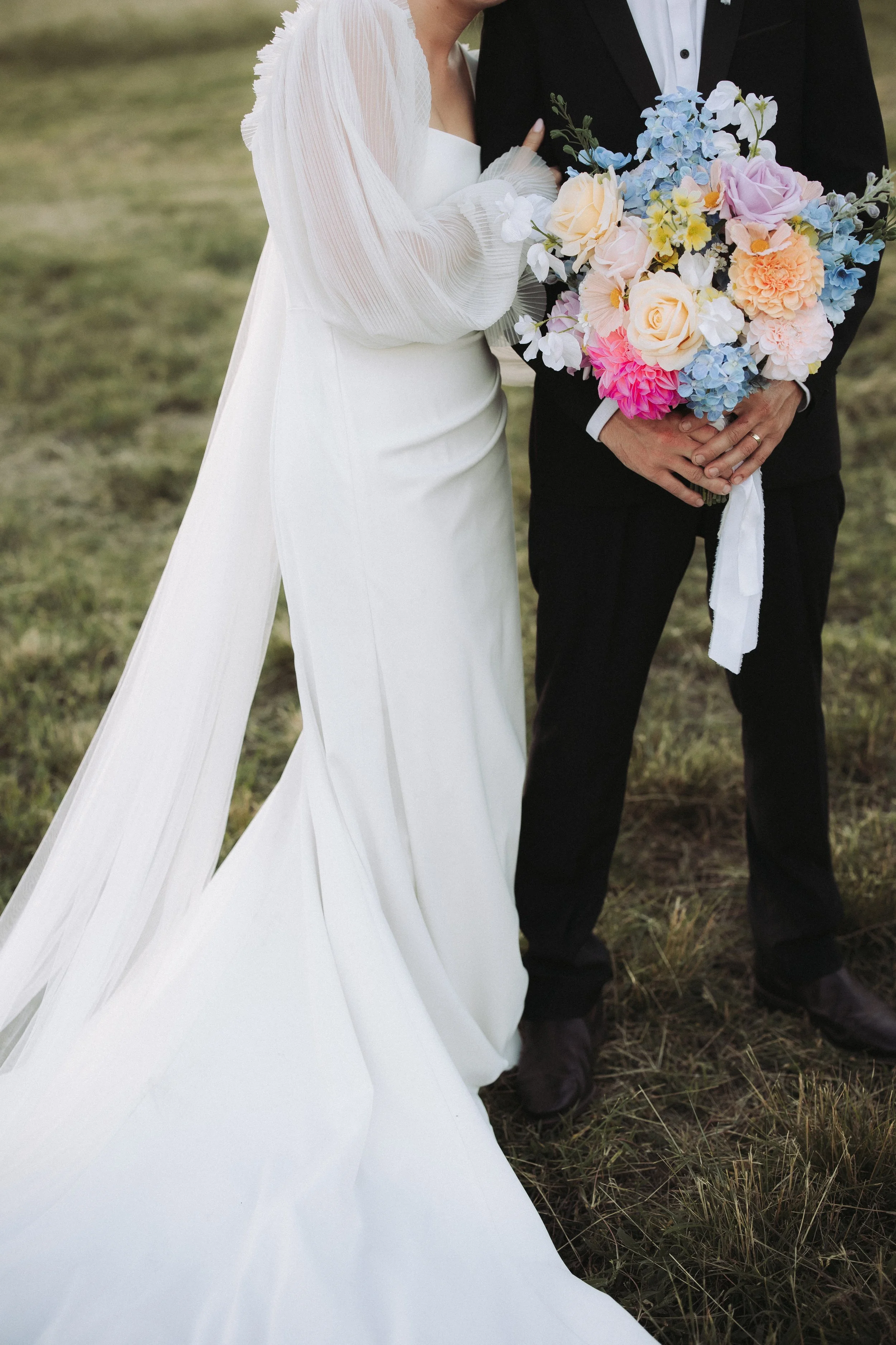 Close-up of a bride and groom standing outdoors, with the bride in a white wedding dress and the groom in a black suit holding a colorful bouquet of flowers.