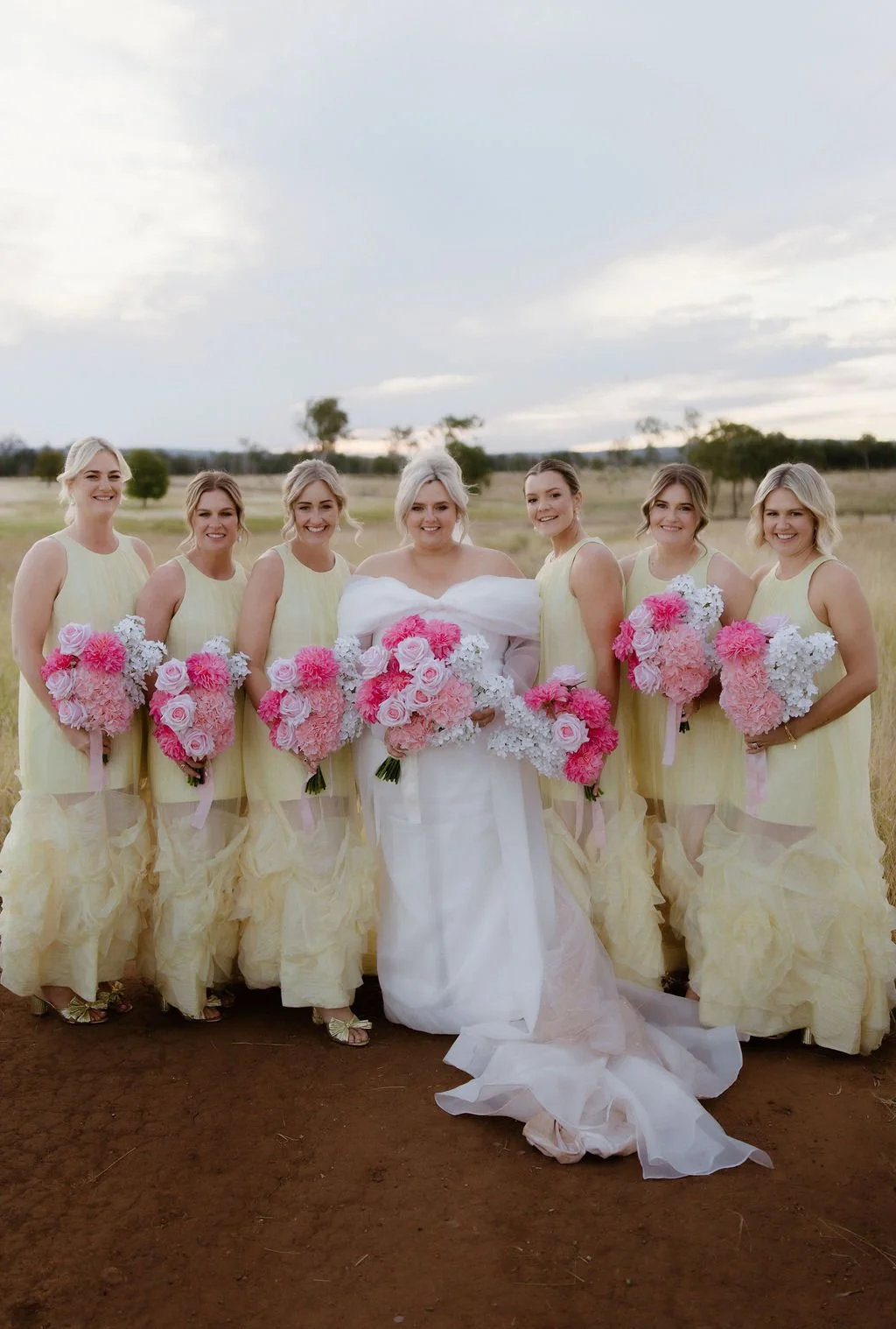 A bride in a white wedding dress and six bridesmaids in light yellow dresses standing outdoors on dirt ground with a field and trees in the background, all holding pink and white floral bouquets.