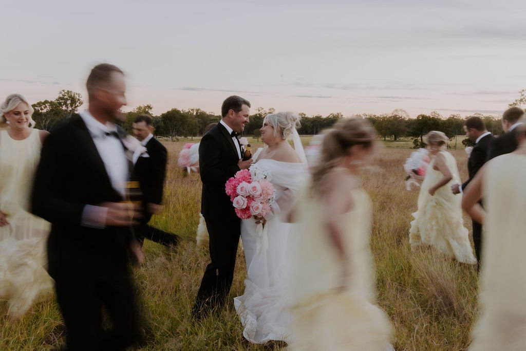 A wedding ceremony in an outdoor field during the evening with the bride and groom in the center, the bride holding a bouquet of pink and white flowers, surrounded by wedding guests in formal attire.