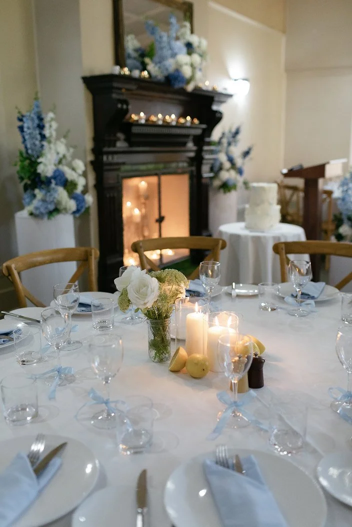 A decorated dining table at an event with a floral centerpiece, candles, and place settings, next to a fireplace with floral arrangements and a cake in the background.