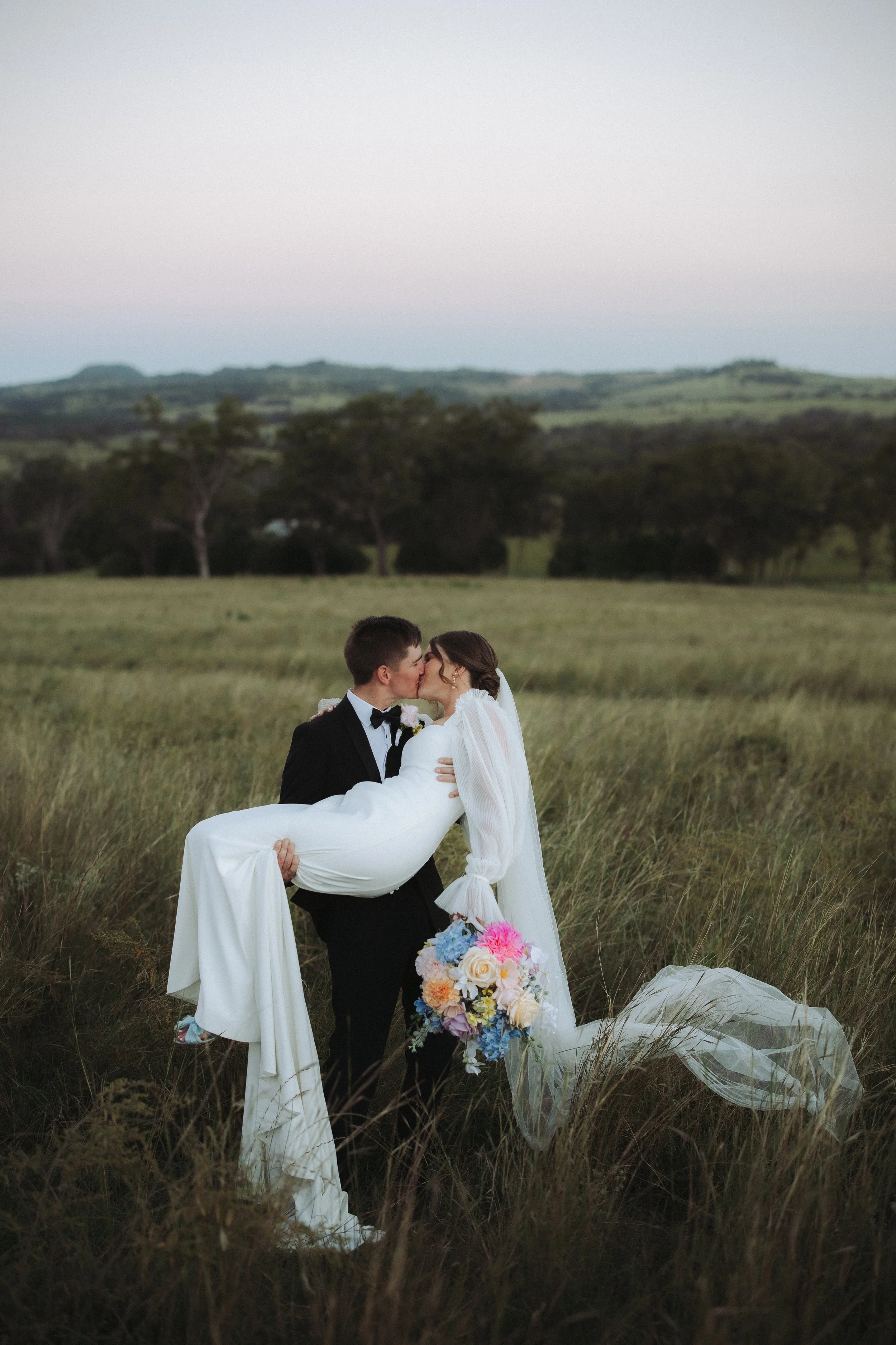 A groom in a tuxedo carries a bride in a white wedding dress through tall grass in an open field with hills and trees in the background.