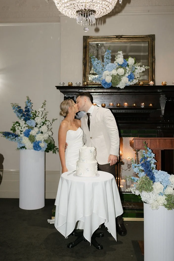 A bride and groom kiss in front of a decorated fireplace with a wedding cake on a small round table. The setting includes large flower arrangements with white and blue flowers, candles, and a crystal chandelier overhead.