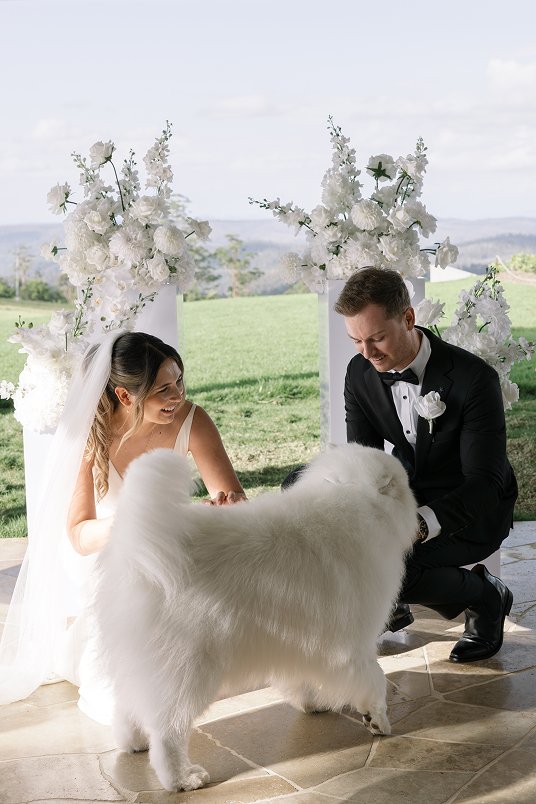 Bride and groom kneeling with a white fluffy dog during wedding ceremony outdoors with floral arrangements and greenery in the background.