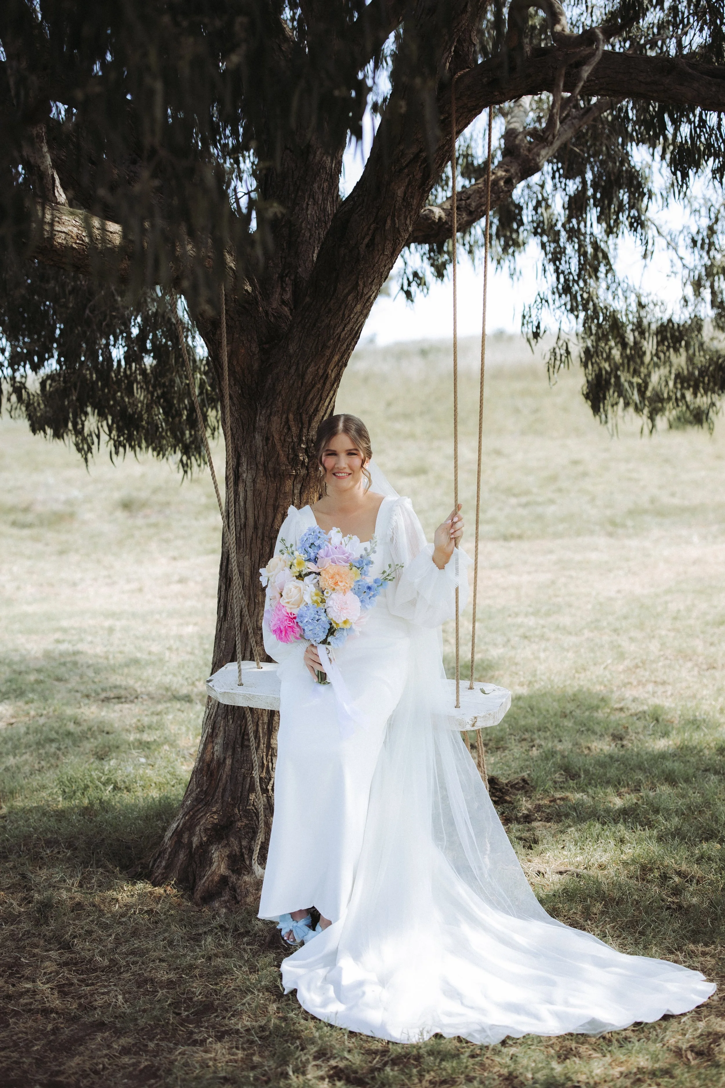 A smiling woman in a white wedding dress sitting on a swing hanging from a tree in an outdoor setting with grass and trees in the background, holding a large bouquet of colorful flowers.