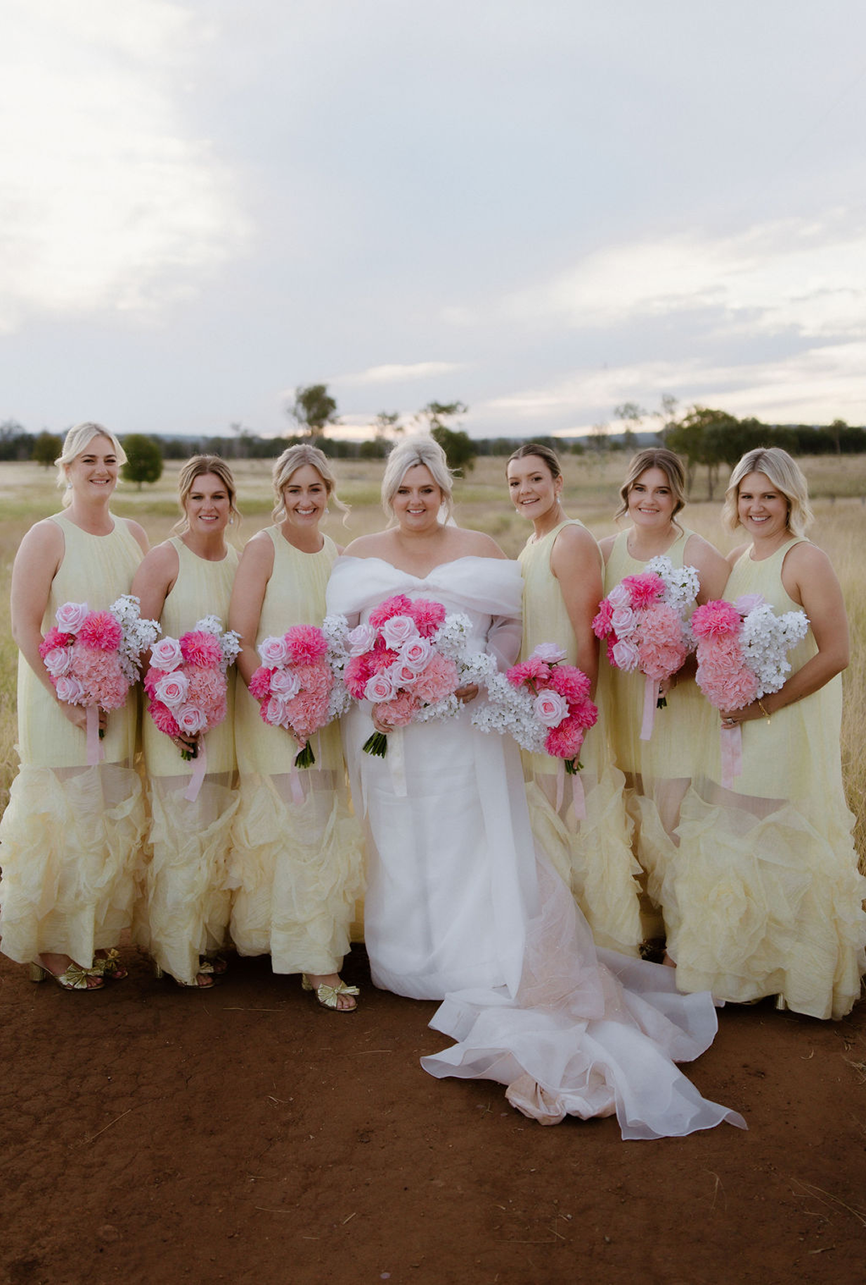 Bridal party with six women in yellow dresses holding pink and white floral bouquets in an outdoor field with a cloudy sky.