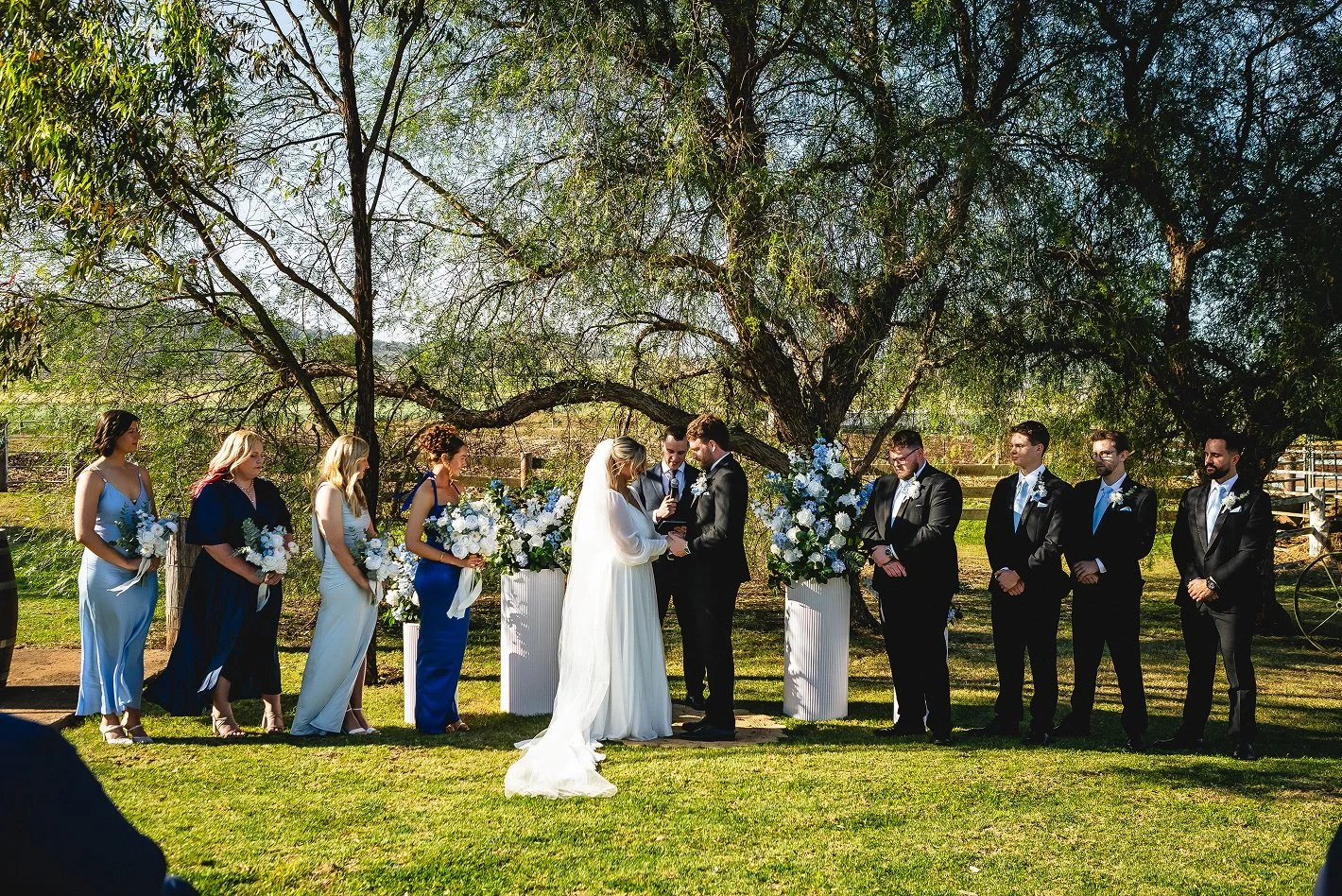 Wedding ceremony outdoors under a large tree with bride and groom exchanging vows surrounded by bridesmaids and groomsmen, with floral arrangements.