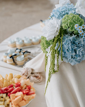 A floral centerpiece with blue hydrangeas, white roses, and green accents on a table with desserts such as assorted cheeses and cupcakes in the background.