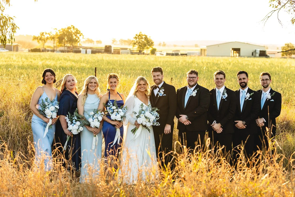 A wedding party standing in a field at sunset, with the bride and groom in the center, surrounded by bridesmaids and groomsmen holding bouquets and dressed in formal attire.