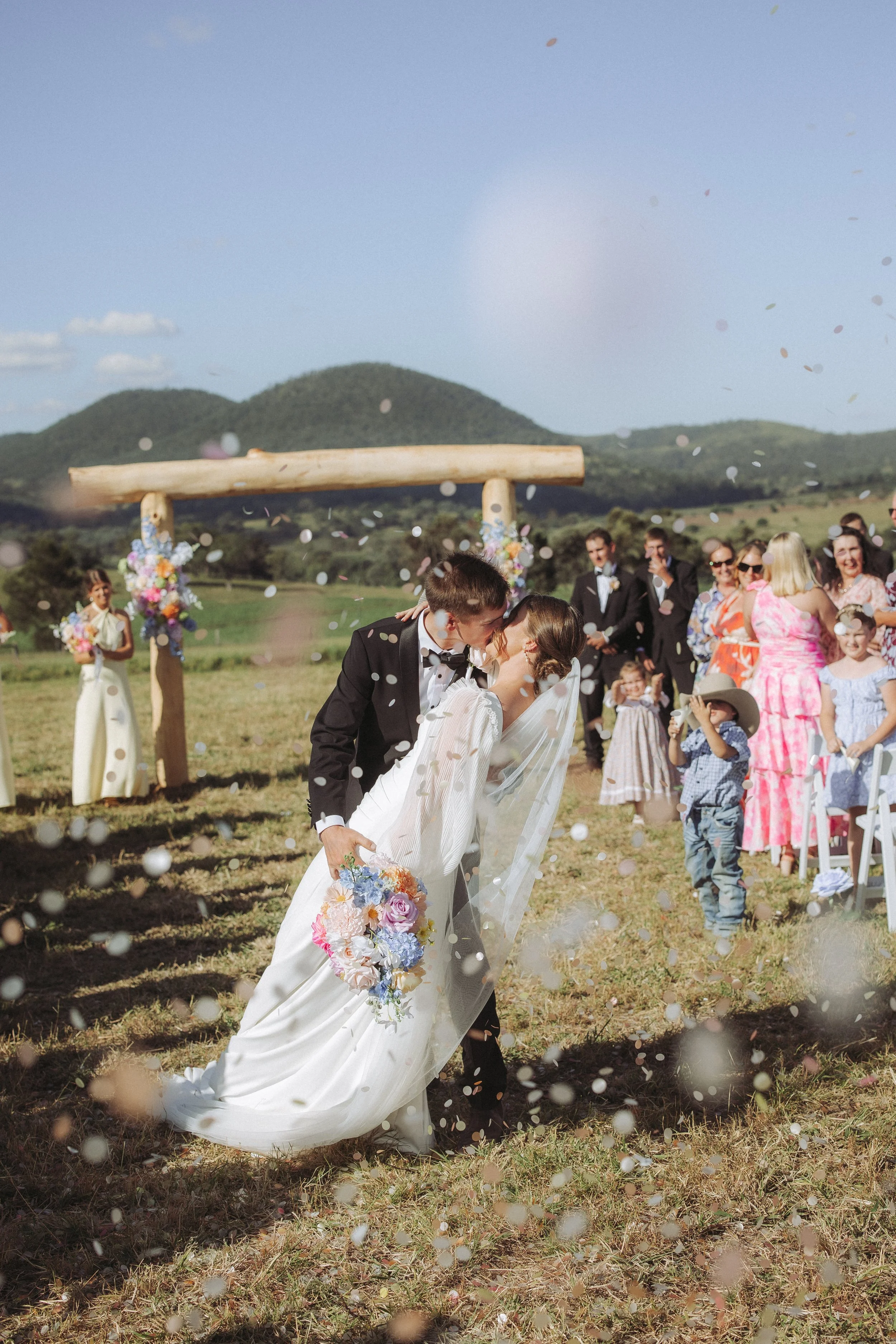 A wedding ceremony outdoors with a bride and groom sharing a kiss, surrounded by guests, mountains, and a wooden arch decorated with flowers. Confetti is falling from above.