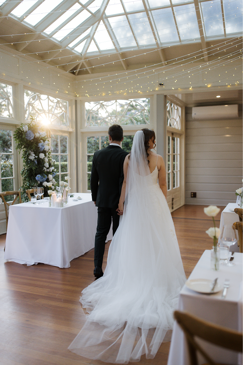 A bride and groom holding hands in a wedding reception hall with large windows, string lights, and floral decorations.