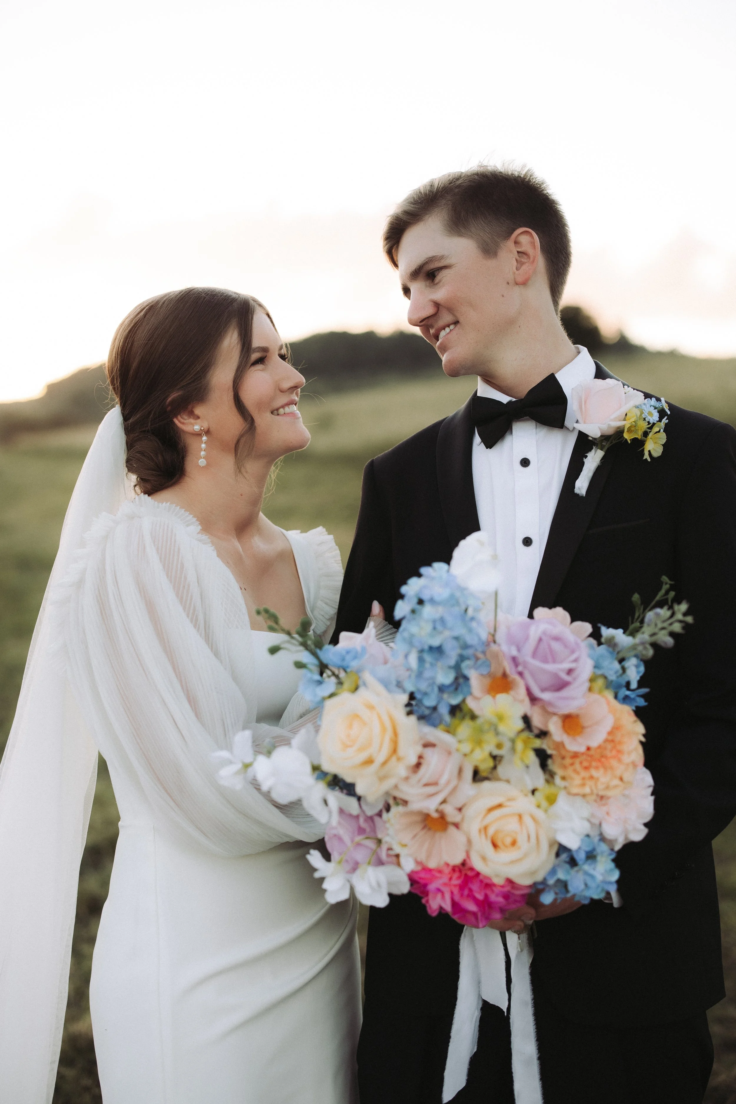 A bride and groom stand close together outdoors during a wedding, smiling at each other, with the bride holding a large bouquet of colorful flowers.