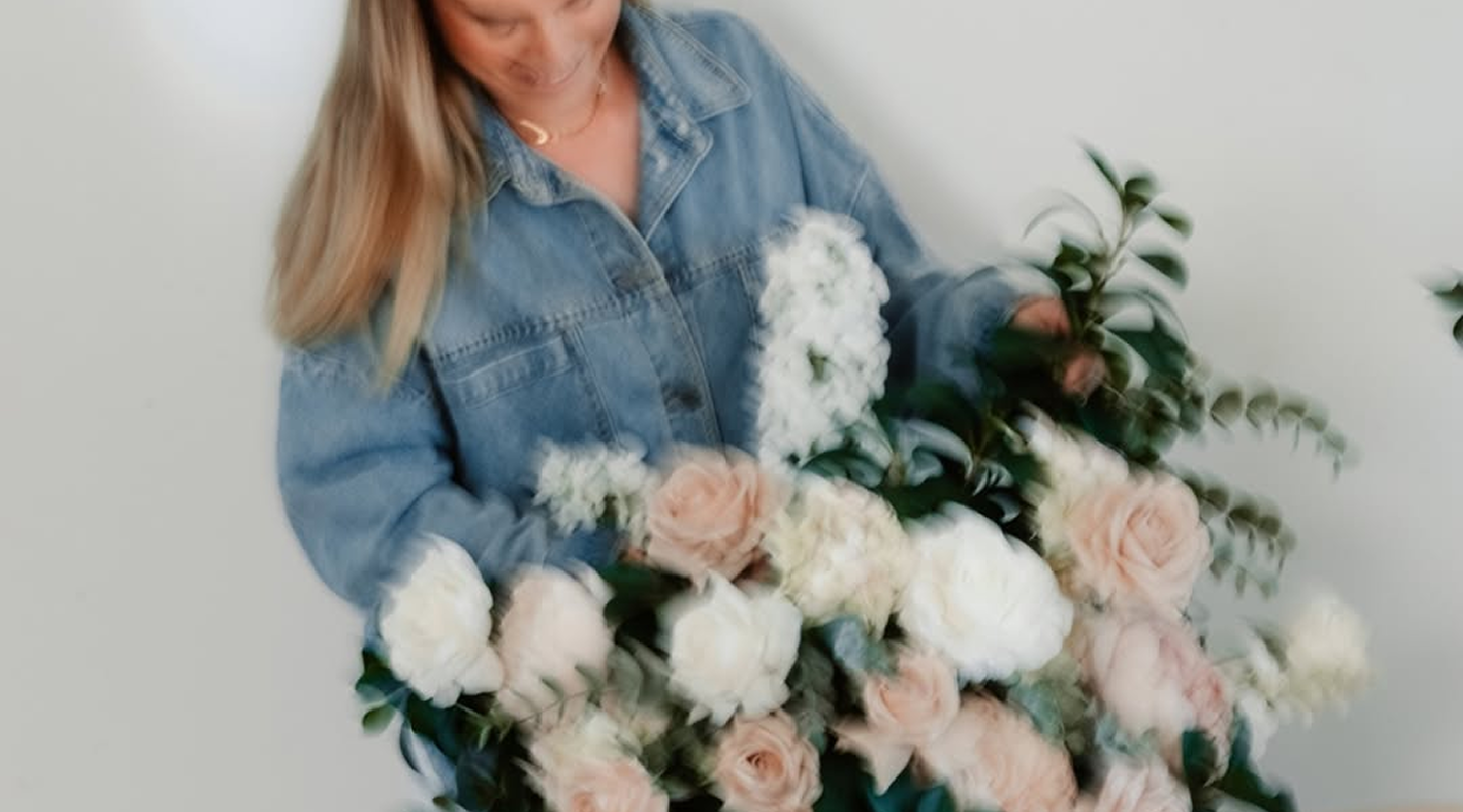 A woman in a denim jacket holding a large bouquet of white and light pink roses and white peonies.