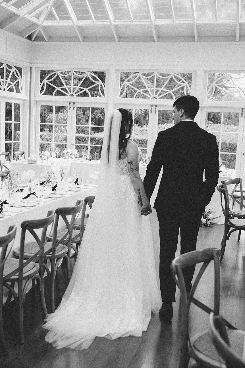A bride and groom holding hands inside a sunroom with large windows, decorated for a wedding celebration.