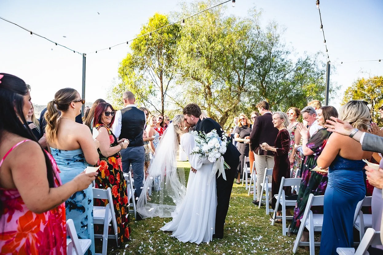 Bride and groom kissing at outdoor wedding ceremony with guests clapping and smiling, under string lights and trees.