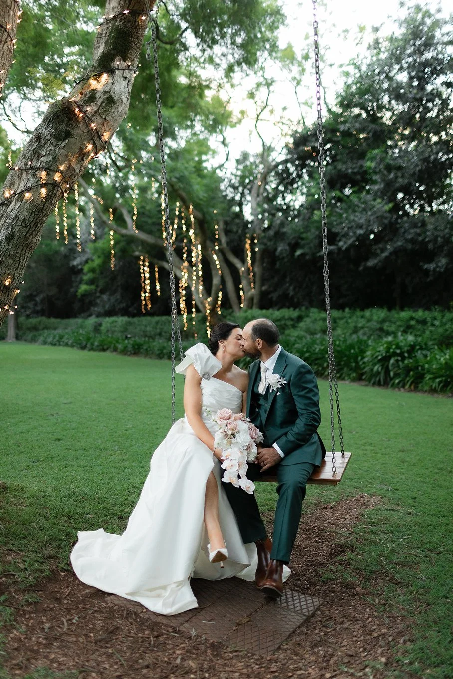 A bride and groom kissing while sitting on a swing hanging from a tree at an outdoor wedding reception during sunset, decorated with string lights.