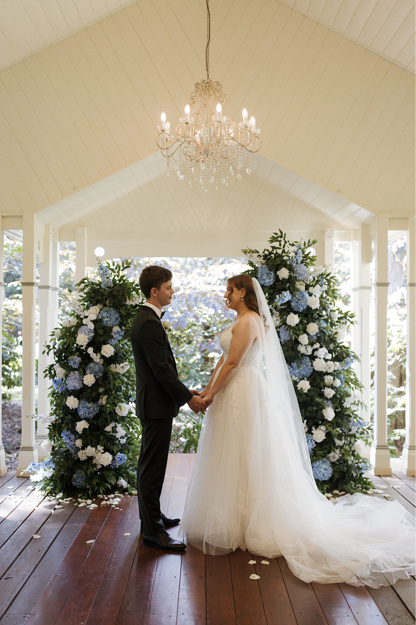 Bride and groom standing face-to-face holding hands inside an decorated gazebo during wedding ceremony, with blue and white floral arrangements behind them, a chandelier hanging from the ceiling, and wooden floorboards.