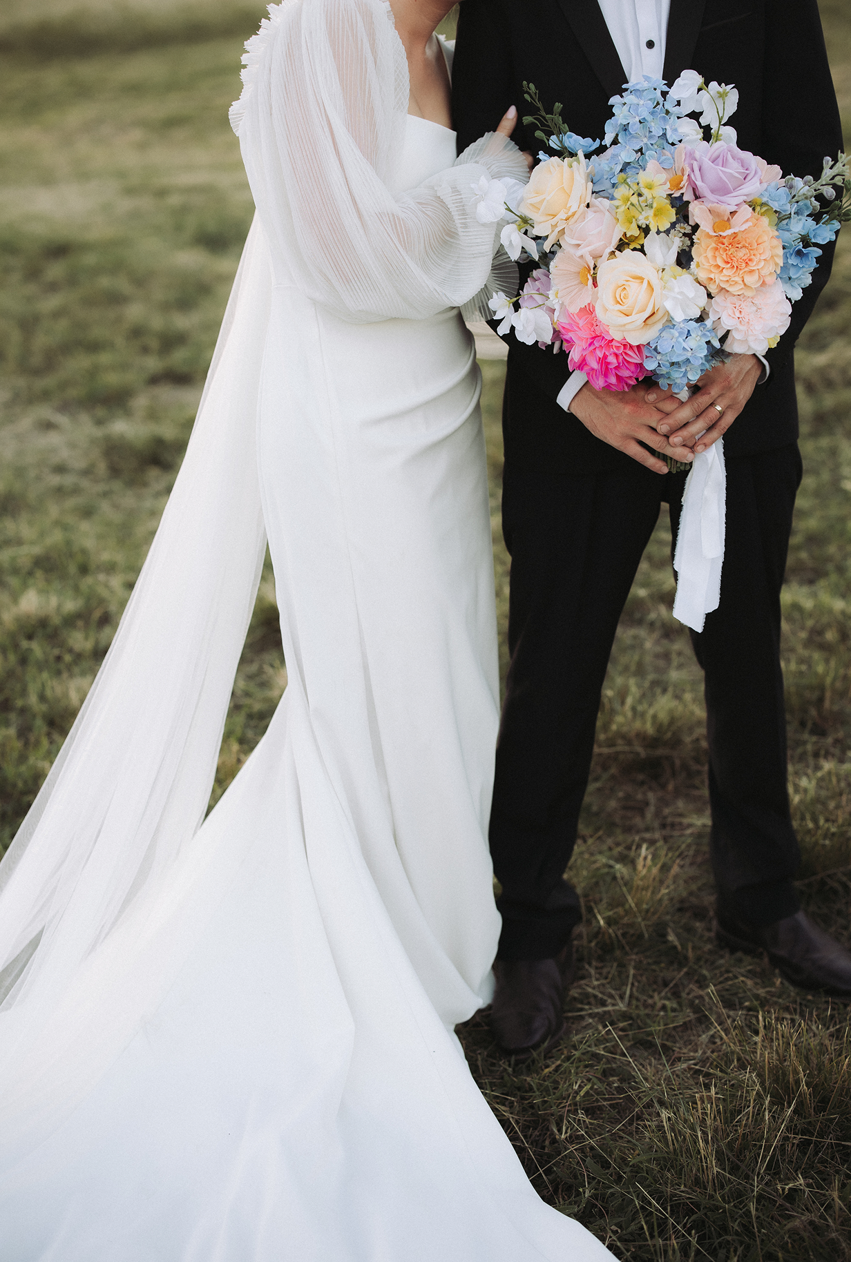 A bride in a white wedding dress and a groom in a black tuxedo holding a colorful bouquet of flowers outdoors on grass.