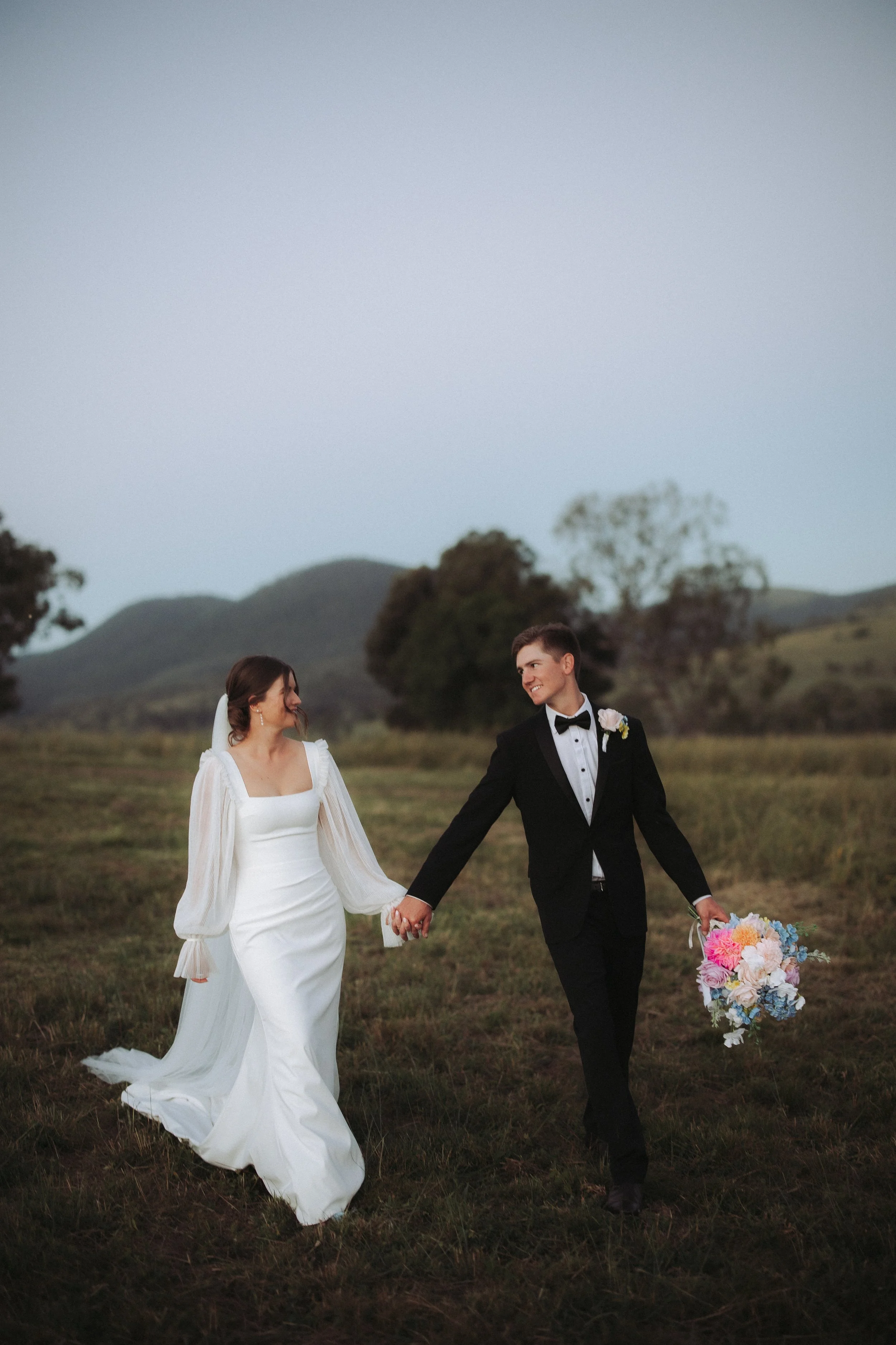 A bride and groom walking hand in hand through a grassy field, smiling at each other, with hills and trees in the background during sunset.