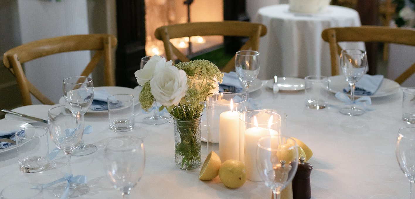 A round dining table set for a formal event with white tablecloth, glassware, napkins, candles, and flowers in a glass jar