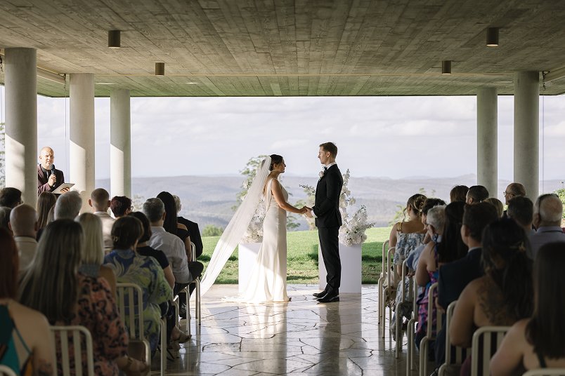 Bride and groom holding hands during wedding ceremony under a pavilion with an open scenic view, officiant on the left, guests seated on either side.