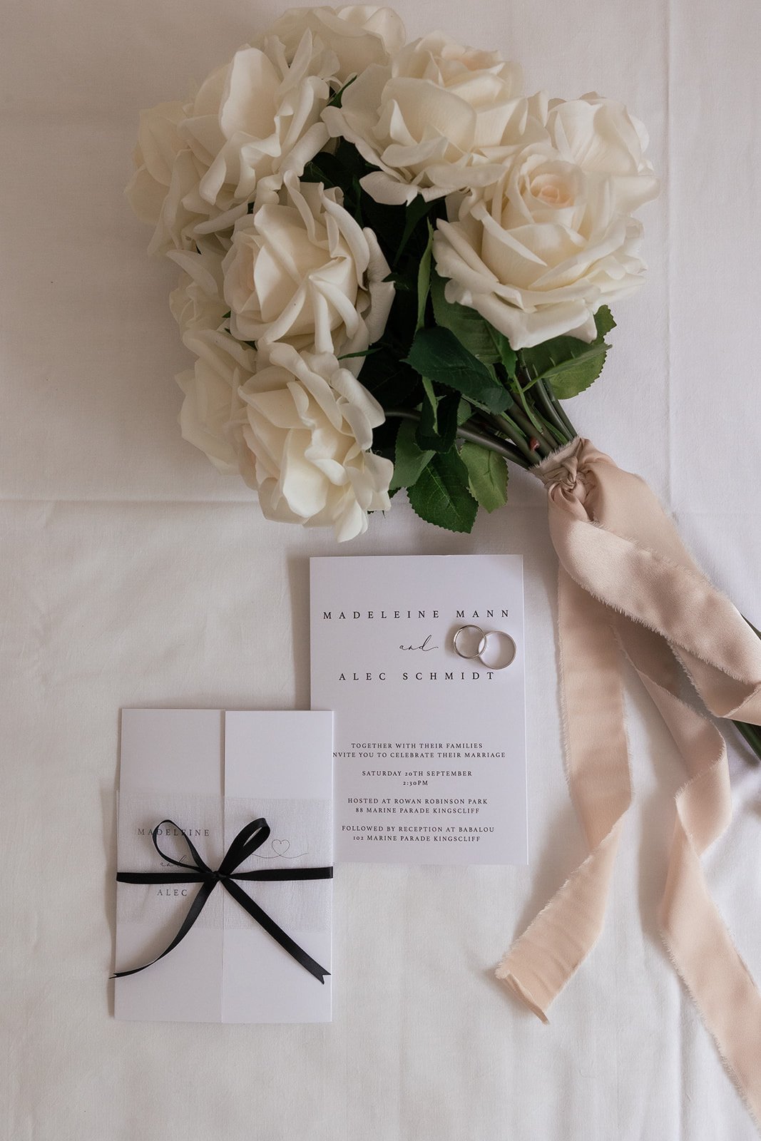A bouquet of white roses with green leaves and a satin ribbon, wedding invitations with rings, and a small wrapped gift with a black ribbon on a white table.