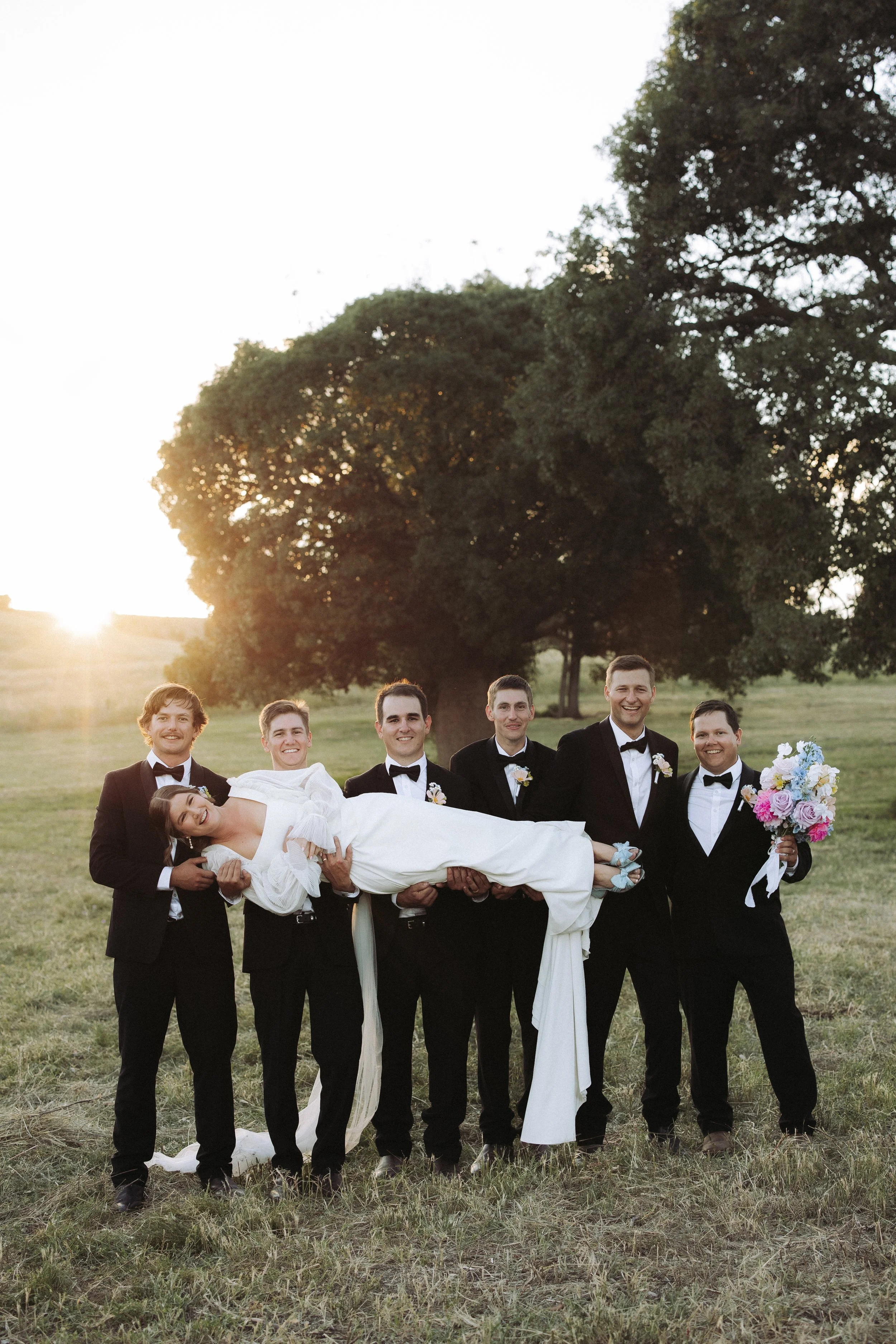 A group of men in tuxedos are holding a bride in a white wedding dress and veil in a field with large trees and the sun setting behind them.