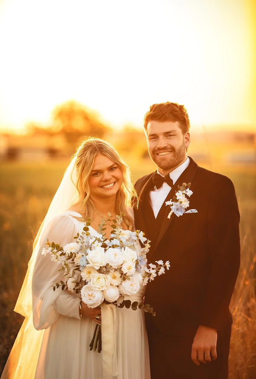 A happy bride and groom standing outdoors at sunset, smiling and holding a bouquet of white flowers. The bride is dressed in a white wedding gown with a veil, and the groom is wearing a black tuxedo with a boutonniere.
