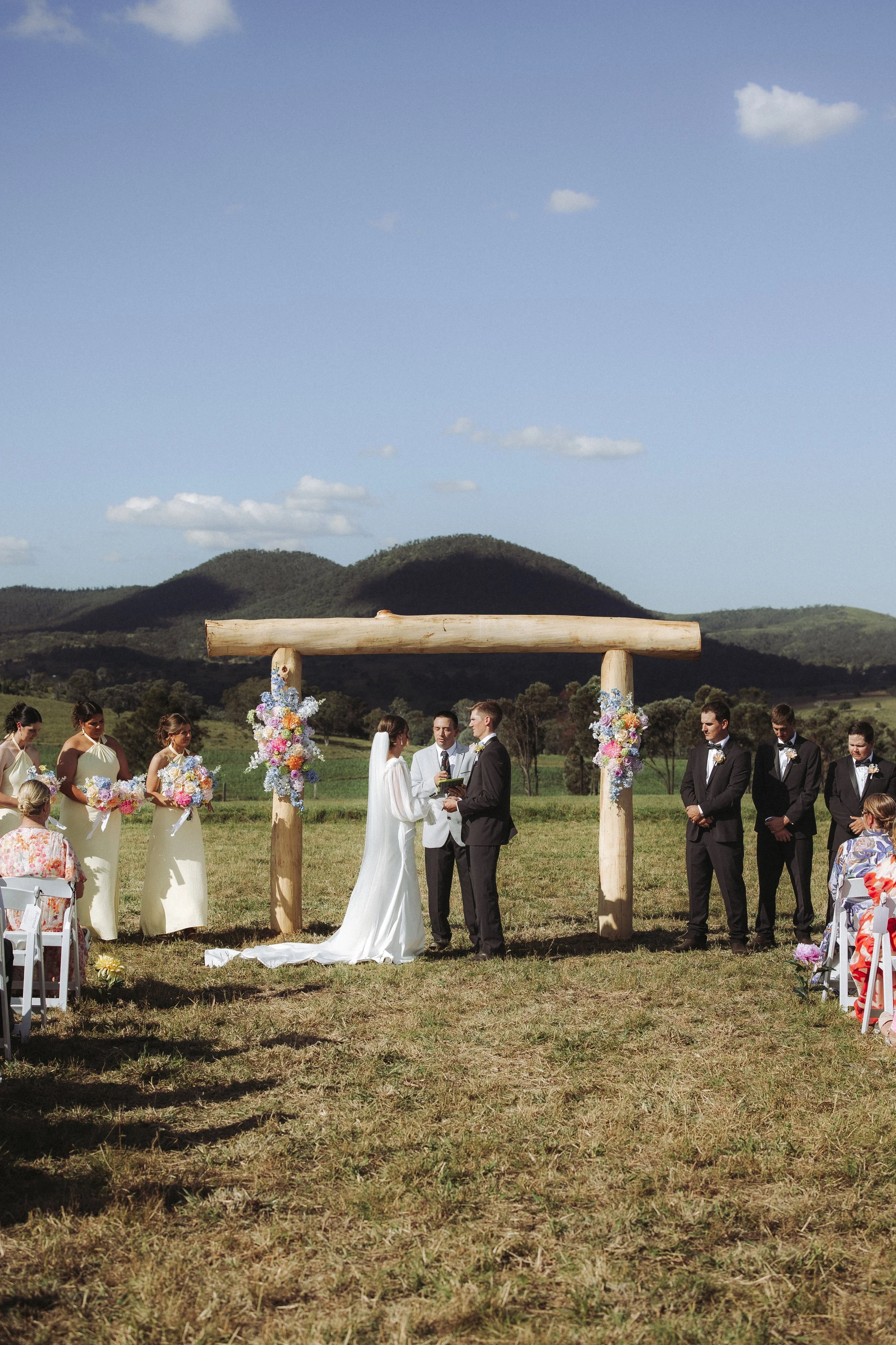 Outdoor wedding ceremony with couple exchanging vows under a wooden arch decorated with colorful flowers, surrounded by bridesmaids and groomsmen, set against hills and blue sky.