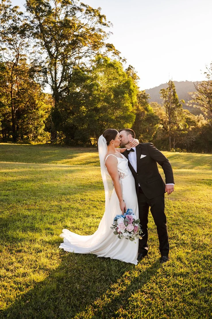 Bride and groom kissing outdoors on a grassy field during sunset, surrounded by trees and mountains, with the bride holding a bouquet of pink and purple flowers.