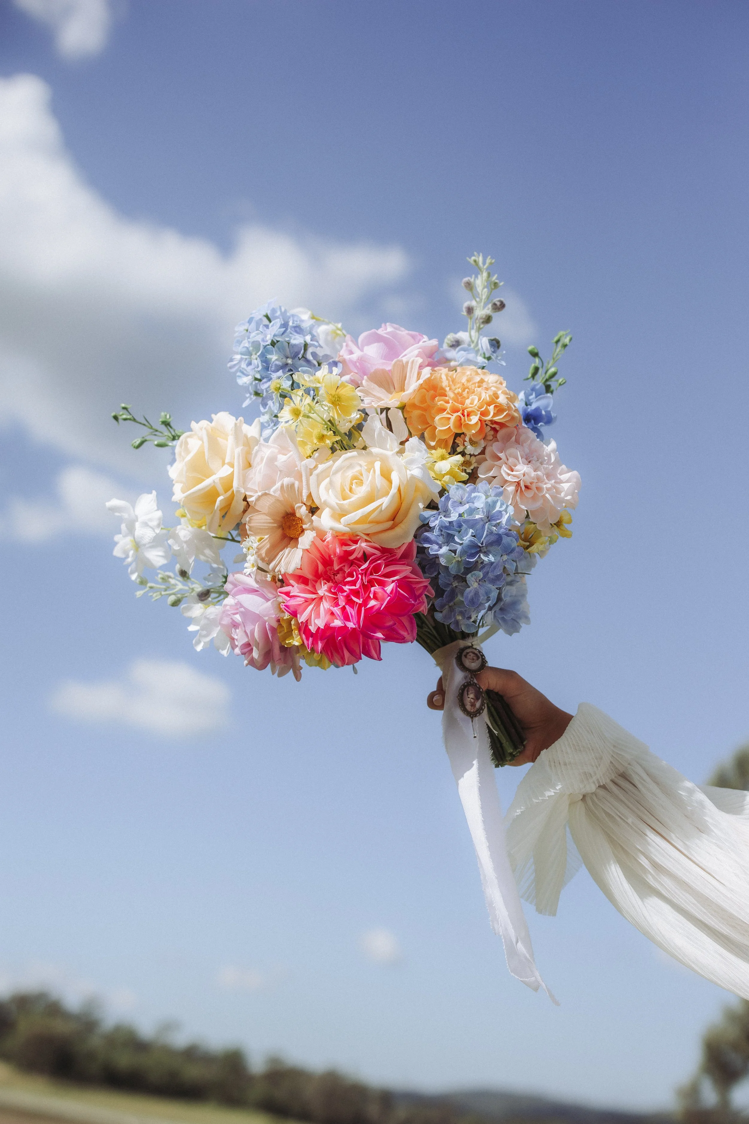 A bouquet of colorful flowers, including roses and hydrangeas, is being held up in front of a clear blue sky with a few clouds.