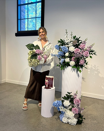 Woman standing next to a large floral arrangement in a white room with a window.