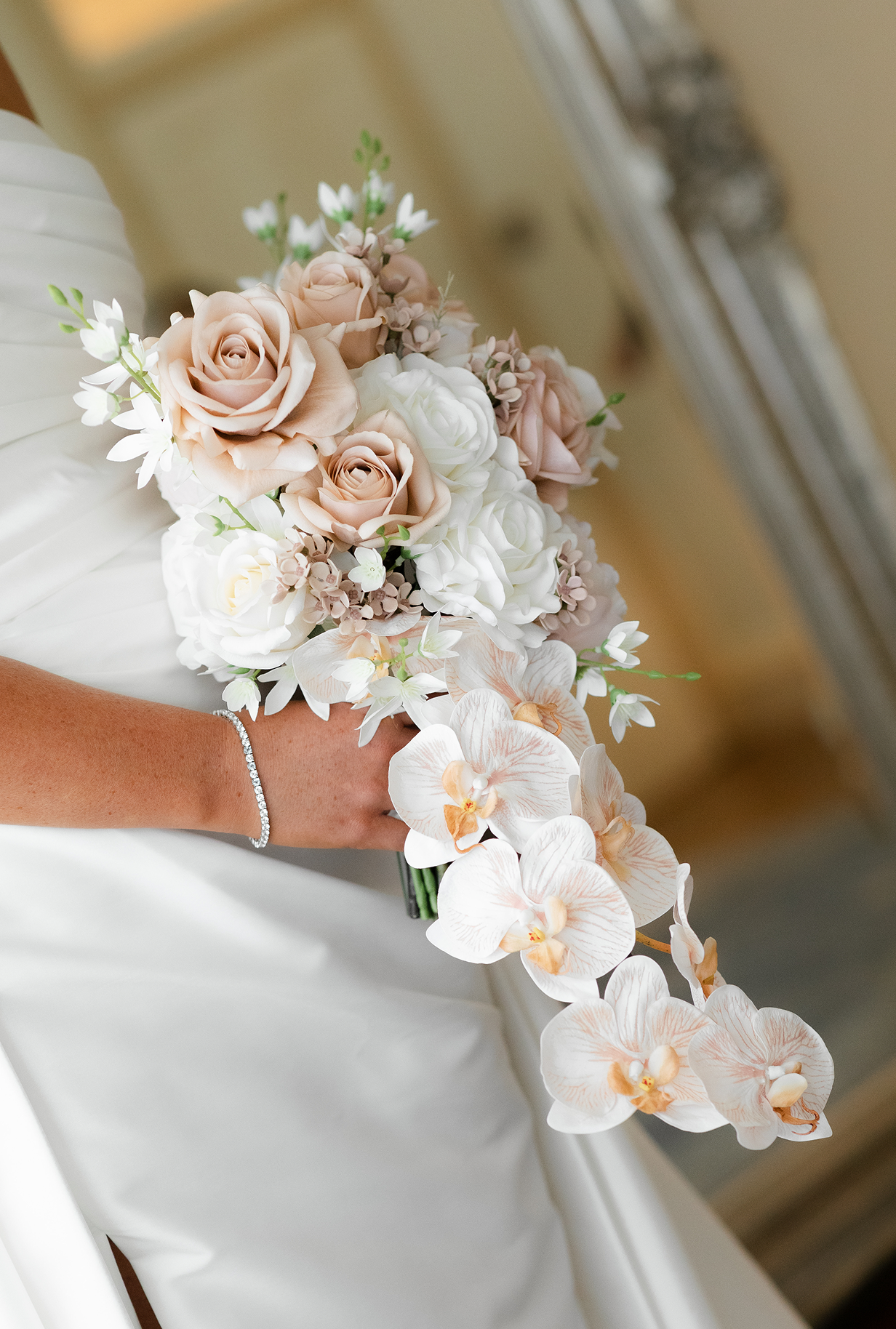 Close-up of a bride holding a cascading bouquet of white and blush pink roses, orchids, and small white flowers, with her hand and a diamond bracelet visible in the foreground.