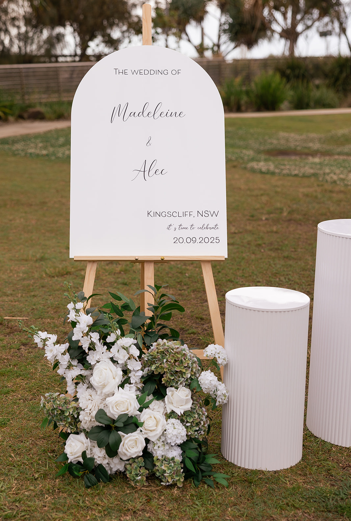 A wedding sign with white background and black elegant font on a wooden easel, surrounded by white flowers and greenery, with two white cylindrical pedestals nearby, set outdoors on a grassy area.