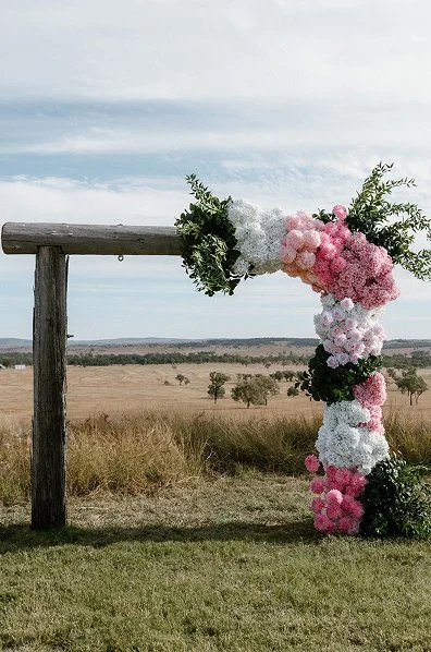 A large floral floral arch with pink, white, and dark flowers, against an open field and cloudy sky.
