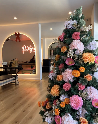 A decorated Christmas tree with pink, peach, and orange flowers and white snow-like accents inside a modern room with a neon sign and furniture.