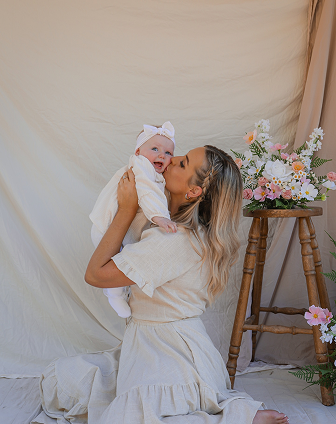 Woman holding a happy baby girl with a bow in her hair, against a cream-colored backdrop with a floral arrangement on a wooden stand.
